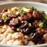 A vibrant plate featuring black beans and fluffy white rice topped with sliced sausage and fresh herbs. The dish is garnished with chopped cilantro and a lime wedge, presented in a rustic bowl on a wooden table.