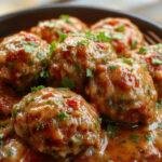 A close-up of a rustic plate featuring golden-brown Italian meatballs generously coated in rich marinara sauce, garnished with fresh basil leaves, with a side of crusty bread and grated Parmesan. The meatballs have a slightly crispy exterior with a moist interior, arranged artfully on the plate.
