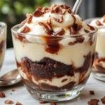 A tall glass layered with rich, creamy cheesecake mixture, fudgy brownie chunks, and a swirl of whipped cream. The parfait showcases distinct layers with a golden-brown brownie base, a smooth white cheesecake layer, topped with cocoa powder and a cherry.
