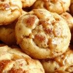 A close-up of golden-brown caramel apple snickerdoodle cookies arranged on a rustic wooden surface. The cookies are topped with a cinnamon sugar coating, with visible caramel bits and apple pieces nestled within. Some cookies are slightly cracked, highlighting their soft texture, and a few cinnamon sticks and apple slices are artfully placed nearby for an inviting autumnal display.
