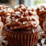 Close-up of moist, chocolatey cupcakes topped with a swirl of glossy marshmallow frosting, sprinkled with cocoa powder, presented on a rustic wooden surface with a spoon and cocoa powder in the background