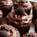 A close-up of a elegantly decorated dark chocolate blackberry cupcake with a glossy blackberries topping, nestled in a gold foil wrapper. The cupcake has a rich, dark chocolate crumb with a smooth chocolate frosting, garnished with fresh blackberries and a light dusting of powdered sugar. The background features a rustic wooden surface with scattered blackberries for a staged, enticing presentation.