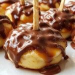 A close-up of multiple caramel apple slices dipped in glossy dark chocolate, arranged on a rustic wooden platter. The vibrant red apple slices are generously coated with smooth, shiny chocolate, with streaks of caramel visible on some edges. The rich contrast between the golden caramel and dark chocolate creates an inviting, indulgent appearance. In the background, blurred autumnal elements evoke a cozy fall atmosphere.