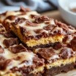 A rich, fudgy brownie square topped with a swirl of creamy pumpkin cheesecake, garnished with a light dusting of cinnamon. The brownies are cut into neat squares, showcasing the marbled topping and moist interior, set on a rustic wooden platter with autumn leaves in the background.