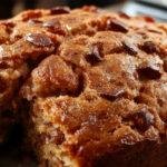 A slice of golden-brown gluten-free banana bread on a white plate, with a slightly cracked top revealing moist interior flecked with banana slices. The bread is garnished with a sprig of fresh mint and a dusting of powdered sugar, set against a rustic wooden background with a light drizzle of honey on the side.