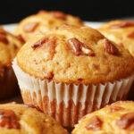 A close-up of golden-brown gluten-free pumpkin muffins topped with a light dusting of powdered sugar, arranged in a rustic basket on a wooden table, with a sprig of fresh cinnamon sticks and pumpkin seeds nearby. The muffins have a moist, spongy texture with visible specks of pumpkin and a slightly cracked top.