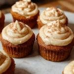 A close-up of three freshly baked pumpkin cupcakes decorated with swirls of creamy frosting, placed on a rustic white plate with a gentle autumn backdrop. The cupcakes have a golden-brown top with hints of orange pumpkin, and the frosting is smooth and inviting, with a slight sheen, topped with a small pumpkin-shaped decoration.