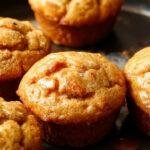 A close-up of a stack of moist, fluffy sweet potato muffins fresh from the oven, golden-brown with a slight crackle on top. The muffins are arranged on a rustic wooden platter with a few crumbs scattered around, highlighting their light, airy texture and appealing color. Soft natural lighting enhances their inviting appearance, with a hint of cinnamon and sweet potato visible in the crumb.