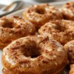 A close-up of three freshly baked pumpkin donuts with a golden-brown exterior, arranged on a rustic white platter. The donuts are dusted lightly with powdered sugar, showcasing their soft, fluffy texture and moist interior. In the background, a small bowl of spiced pumpkin glaze and a few cinnamon sticks are visible, adding warm autumnal accents to the scene.
