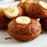 Golden-brown banana donut holes with a slightly crispy exterior and soft interior, arranged on a white platter with a few crumbs scattered around, garnished with a light dusting of cinnamon and sliced bananas in the background.