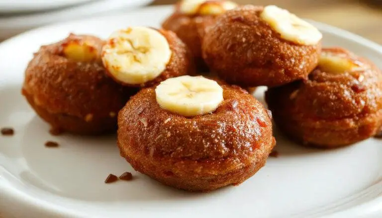Golden-brown banana donut holes with a slightly crispy exterior and soft interior, arranged on a white platter with a few crumbs scattered around, garnished with a light dusting of cinnamon and sliced bananas in the background.