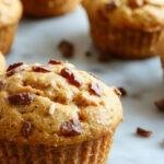 A close-up of a batch of golden-brown bakery-style pumpkin muffins arranged on a rustic wooden platter, topped with a light dusting of powdered sugar and hints of cinnamon. The muffins have a tender crumb with a slight rise and are presented with a few fresh pumpkin seeds and a drizzle of caramel sauce for added appeal.