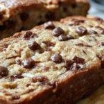 A golden-brown banana bread loaf topped with semi-sweet chocolate chips, sliced to reveal a moist crumb with chunks of bananas and chocolate chips visible inside; the loaf rests on a rustic wooden surface with a few chocolate chips scattered around, evoking a warm, inviting atmosphere.