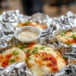 A close-up of a foil packet with grilled vegetables and chunks of meat, golden and slightly charred edges, placed on a rustic wooden surface with a background of a campfire setting.