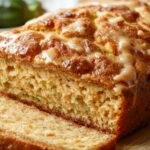 A golden-brown zucchini bread loaf with a moist texture, topped with a light glaze and sliced to reveal a tender crumb dotted with green zucchini pieces. The bread sits on a rustic wooden surface with a few shredded zucchini and a teaspoon of cinnamon nearby, highlighting its fresh and inviting appearance.