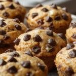A close-up of golden-brown pumpkin muffins with chocolate chips scattered on top, arranged on a rustic wooden tray. The muffins have a moist, fluffy texture with slightly cracked tops and melty chocolate chips visible. A few muffins are stacked, showcasing the soft crumb inside, and there is a hint of cinnamon and pumpkin in the background for added warmth.