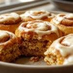 A close-up of freshly baked pumpkin cinnamon rolls topped with smooth, white cream cheese frosting, arranged on a rustic wooden platter. The golden-brown rolls are drizzled with more frosting, revealing swirls of pumpkin and cinnamon inside. The background features a soft autumnal setting with hints of cinnamon sticks and pumpkin decorations, emphasizing warmth and comfort.