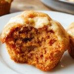 A close-up of golden-brown pumpkin cream cheese muffins arranged on a rustic wooden platter. The muffins are topped with a swirl of cream cheese filling, slightly melted, with a few cinnamon sugar sprinkled on top. The background includes a cozy fall setting with mini pumpkins, cinnamon sticks, and a tartan cloth, enhancing the warm, inviting appearance.