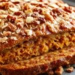 A golden-brown pumpkin loaf topped with a crumbly streusel streusel topping, sliced to reveal moist orange interior with specks of spice, served on a rustic wooden board with a fork and a cup of coffee in the background.