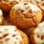 A close-up of a plate of festive pumpkin sugar cookies topped with smooth, white cream cheese frosting. The cookies have a golden-brown hue with a textured surface, garnished with tiny specks of cinnamon and a drizzle of glaze. The background features a rustic wooden table with autumn leaves, enhancing the seasonal feel.