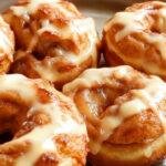 Golden-brown puff pastry doughnuts filled with sliced apple pieces, topped with a light dusting of powdered sugar and arranged on a white plate with a rustic cloth background, capturing their flaky, crispy texture and appealing interior filling.