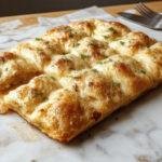 Une focaccia maison ultra moelleuse dorée, garnie de gros sel, herbes fraîches et un filet d'huile d'olive, présentée sur une planche en bois rustique.
