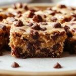 A close-up of rectangular healthy no-bake cookie dough bars placed on a rustic wooden serving board. The bars have a golden-brown crust with visible chocolate chips and a creamy, slightly textured surface. Fresh mint leaves and a drizzle of dark chocolate are artfully arranged around the bars, highlighting their indulgent yet healthy appeal.
