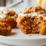 A batch of golden-brown pumpkin muffins topped with a sprinkle of chopped pecans and a drizzle of honey, arranged on a rustic wooden platter with a few pumpkin seeds scattered around. The muffins are varied in size, with some slightly cracked surfaces revealing a moist interior, accompanied by a small bowl of Greek yogurt on the side.