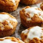 Golden-brown pumpkin muffins topped with a light dusting of cinnamon, arranged on a rustic wooden platter with a fork beside them. The muffins reveal a moist, textured crumb with visible specks of pumpkin and a hint of creamy Greek yogurt swirled into the batter, creating an inviting autumnal display.