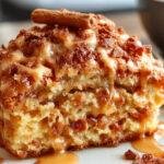 A close-up of a slice of apple crumb cake with a golden-brown crumb topping, drizzled with cinnamon glaze, and garnished with a sprinkle of cinnamon. The cake sits on a rustic wooden platter with visible apple chunks inside, steam gently rising from the warm dessert.