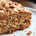 A freshly baked cinnamon crunch banana bread loaf with a golden-brown crust topped with crunchy cinnamon sugar crumbs. The bread is sliced to reveal a moist, tender interior dotted with ripe banana chunks and a hint of cinnamon. The loaf rests on a rustic wooden surface, surrounded by scattered cinnamon sticks and a dusting of powdered sugar, highlighting its warm, inviting appearance.