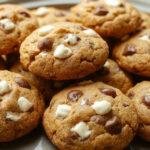 A close-up of a freshly baked pumpkin white chocolate chip cookie on a rustic wooden surface. The cookie is golden-brown with visible white chocolate chips and a slightly cracked top revealing a soft interior. Surrounding the cookie are hints of cinnamon and nutmeg, with a few white chocolate chips scattered nearby, emphasizing the warm fall theme.