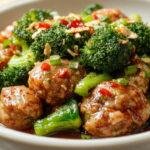 A vibrant plate featuring glossy Mongolian meatballs glazed in a savory sauce, nestled among bright green broccoli florets. The meatballs are round and richly colored, with a slightly caramelized exterior, and are arranged neatly with some sauce drizzling over them. The broccoli adds a fresh, crisp contrast, and the dish is presented on a modern white plate with a clean, minimalist background revealing a hint of chopsticks for added Asian-inspired flair.