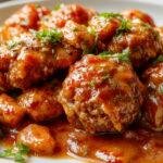 A close-up of perfectly round, golden-brown ground beef meatballs arranged on a white plate, topped with fresh parsley and served with a side of spaghetti in a rich tomato sauce. The meatballs have a crispy exterior with visible herbs and juiciness inside, creating a mouthwatering presentation.