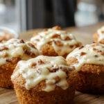 A batch of golden-brown bakery-style pumpkin cheesecake muffins arranged on a rustic wooden tray. The muffins have a slightly cracked top, revealing creamy pumpkin cheesecake swirls inside. Some muffins show a dollop of cream cheese filling peeking through, with a light dusting of cinnamon on top. The background features a lightly blurred autumn-themed backdrop with warm, cozy tones.