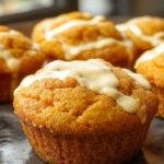 A close-up of a freshly baked pumpkin cream cheese muffin with a golden-brown top, swirls of cream cheese filling visible from the side, subtle fall-themed garnishes like cinnamon stick and a sprig of thyme on a rustic white plate