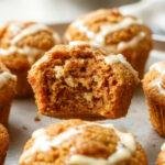 Golden-brown pumpkin cream cheese muffins arranged in a rustic wire basket lined with a parchment paper liner. The muffins have a slightly domed top with a swirl of cream cheese visible through cracks in the crust, and a soft, moist crumb texture with hints of orange pumpkin color. Some muffins are partially broken open to reveal the creamy filling inside, with a few crumbs scattered around creating an inviting, homemade feel.