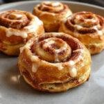 A close-up of pumpkin-shaped cinnamon rolls topped with creamy white frosting, arranged beautifully on a rustic wooden platter with a sprinkle of cinnamon on top. The rolls are golden-brown with visible swirls of pumpkin and cinnamon filling, glistening with a slight glaze and dusted with powdered sugar around the edges.