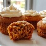 A close-up of two fluffy pumpkin cupcakes topped with generous swirls of creamy white cream cheese frosting, decorated with a sprinkle of cinnamon on top, presented on a rustic wooden plate with a blurred autumnal background.