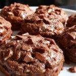 A close-up of three golden-brown chocolate biscuits arranged on a white plate with a drizzle of melted chocolate on top, surrounded by cocoa powder and chocolate shavings, with a slightly blurred background of a baking sheet and ingredients.
