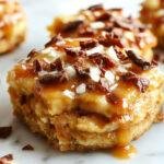 A close-up of salted caramel apple bars arranged on a rustic wooden platter. The bars have a golden-brown crust topped with caramel sauce and sliced fresh apples with a sprinkle of sea salt. The caramel glistens, and the apples are vibrant red and green, contrasting beautifully with the rich caramel and flaky crust. Light crumbs are scattered around, highlighting their homemade appeal.