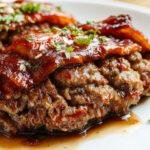 A golden-brown bacon-wrapped meatloaf sliced open to reveal a juicy, tender interior. The bacon lattice on top crisps beautifully around the edges, garnished with fresh herbs. The dish is plated on a rustic wooden surface with a side of green vegetables, showcasing a homey yet elegant presentation.