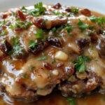 A close-up of a plated Salisbury Steak featuring two browned beef patties topped with a glossy, dark brown rich gravy. The steaks are garnished with a sprig of fresh parsley and served alongside a portion of buttery mashed potatoes and steamed green beans. The presentation is on a white ceramic plate, with the gravy perfectly pooling around the patties, highlighting their tender textures and savory appearance.