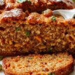 A golden-brown loaf of zucchini carrot bread sliced to reveal a moist interior flecked with grated zucchini and carrots, served on a rustic wooden cutting board with a sprig of fresh herbs on top.