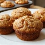 A close-up of golden-brown pumpkin muffins arranged on a rustic wooden plate, with some muffins topped with a light sprinkle of cinnamon and a slice of pumpkin on the side. The muffins display a moist, dense texture with a slightly cracked surface, capturing their warm, inviting appearance.