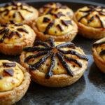 A set of four small, spooky Halloween-themed mini pies arranged on a dark black plate. Each pie features a jagged crust edge and creepy toppings like icing eyes, spider web designs, and pumpkin faces. The pies have a golden-brown crust with colorful themed decorations in orange, black, and white, creating a festive, spooky appearance against a black background with Halloween-themed props in the scene.