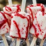 A close-up of three Blood Splatter Cakesicles arranged on a dark plate. The cakesicles are coated in glossy red icing with realistic blood splatter effects and black edible glitter accents, giving a spooky and eerie appearance. They are decorated with small plastic Halloween-themed toppers and are set against a black background, emphasizing their creepy yet sweet presentation.