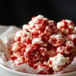 A close-up of vibrant red velvet popcorn with a glossy coating, presented in a spooky-themed bowl with occasional black and orange accents, highlighting the popcorn's rich red color and textured surface, creating a festive Halloween vibe.