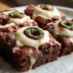 A close-up of spooky eyeball brownies arranged on a dark serving platter. Each brownie is topped with a white glazed icing, featuring a red and black candy eye in the center, creating a creepy yet tempting appearance. The textures showcase a fudgy bottom and smooth icing, with the eyes giving a realistic eyeball effect. The background is dark, enhancing the eerie Halloween theme.