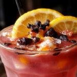 A tall glass of vibrant yellow Witches Brew Lemonade garnished with a spooky rubber spider and a slice of lemon on the rim. The drink is served over ice in a clear glass, with a spooky Halloween themed background featuring cobwebs and orange-tinted lighting, creating a festive and eerie atmosphere.